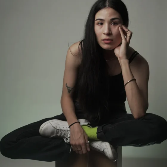 A woman with long, straight, black hair, sitting criss-cross on a stool and resting her head in her left hand. She sits in front of a gray background, and wears a sleeveless, black top with black pants, white shoes, and yellow socks.