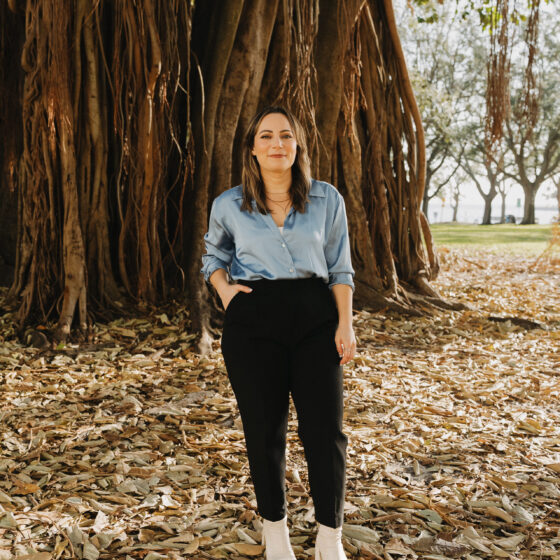 A woman with long, straight, brown hair, standing in front of a giant tree trunk and fallen leaves, with her right hand in her pocket. She wears a silky, blue button-down, black pants, and white shoes.