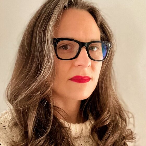 A woman with long, wavy, brown-and-gray hair. She stands in front of a beige wall, and wears a white sweater and black, rectangular glasses.