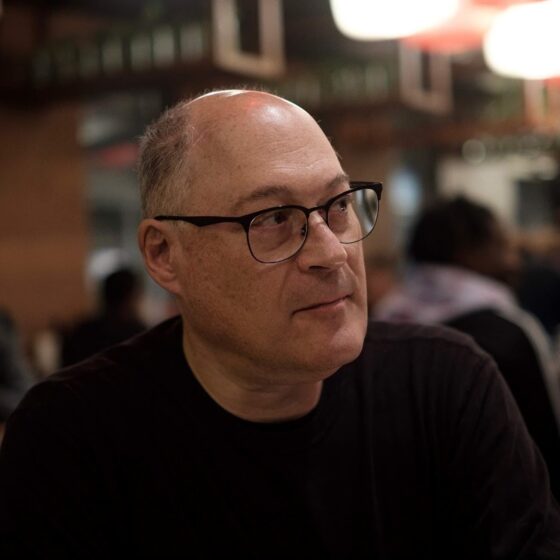 A bald man, looking to the right. He is sitting against the background of a blurred restaurant and wearing a black shirt and rectangular glasses.