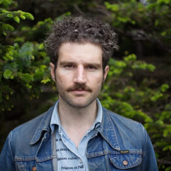 A man with curly, brown hair and a mustache. He stands outside in a wooded area, and wears a blue button-up with back text all over it underneath a blue jean jacket.