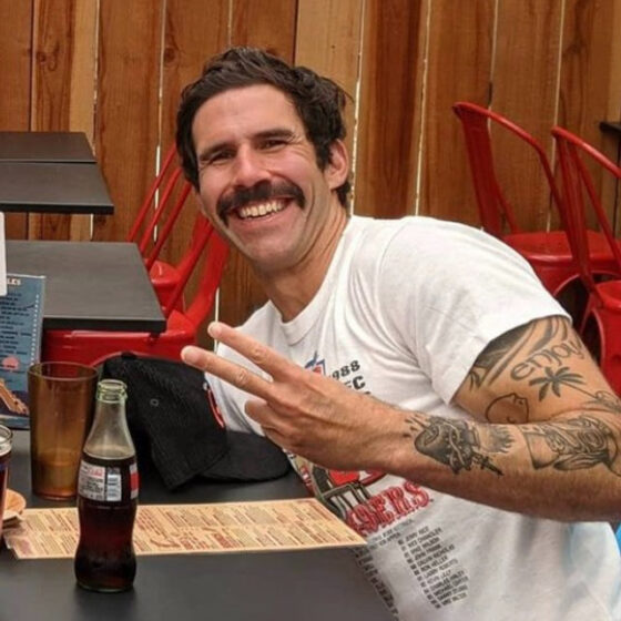 A man with short, straight, brown hair, a large mustache, and brown eyes. He is smiling and bent toward the left, flashing a peace sign with his left arm. He is in some sort of restaurant with wooden walls, black-and-red tables, and red chairs. The table he is sitting at has a glass bottle full of Coca-Cola. on it. He wears a white T-shirt.