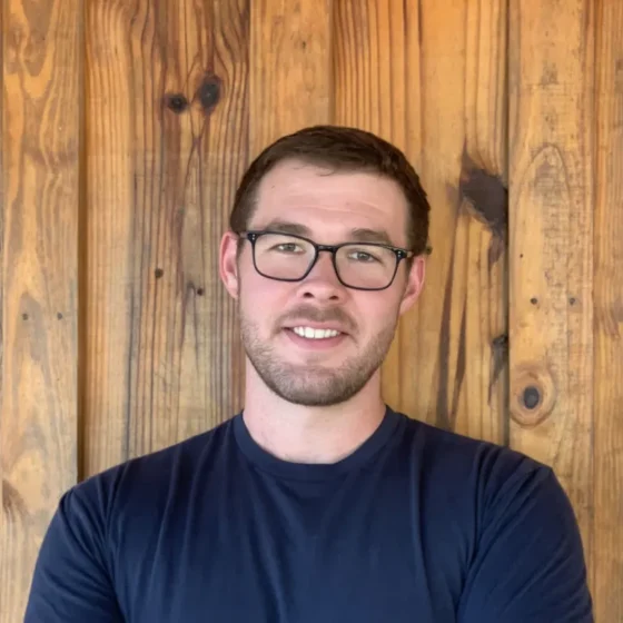A man with buzzed, brown hair, a short beard, and brown eyes, smiling. He stands in front of a wooden wall, and wears a navy T-shirt and squared-shaped glasses.
