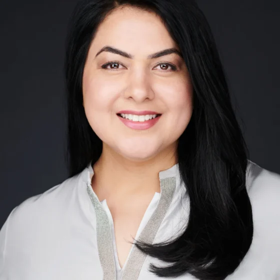 A woman with straight, black hair that goes past her shoulders, smiling. She is standing in front of a black background and wearing a collared, white shirt with silver detailing.