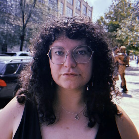 A woman with long, curly, brown hair and bangs. She is standing outside on a city sidewalk, wearing a black tank-top, a silver necklace, and hexagonal glasses.