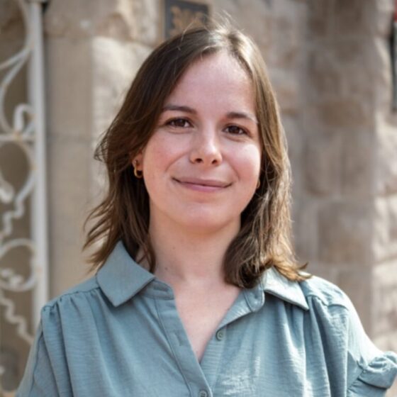 A woman with straight, brown hair that goes down to her shoulders, smiling. She stands outside a stone building, and wears a teal-blue, button-up T-shirt.