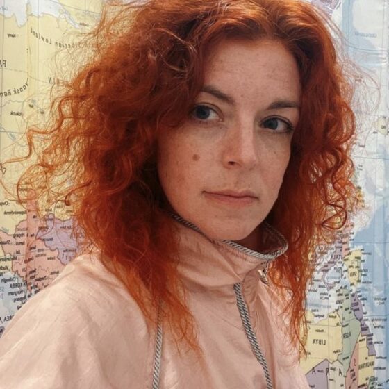 A woman with curly, red hair that goes past her shoulders and dark eyes. She stands in front of a world map background, and wears a pink quarter-zip.