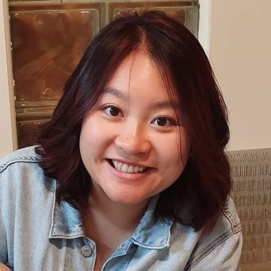 A woman with shoulder-length, straight, black hair, smiling. She stands in front of a wooden-paneled door on a white wall, and wears a light-jean shirt.