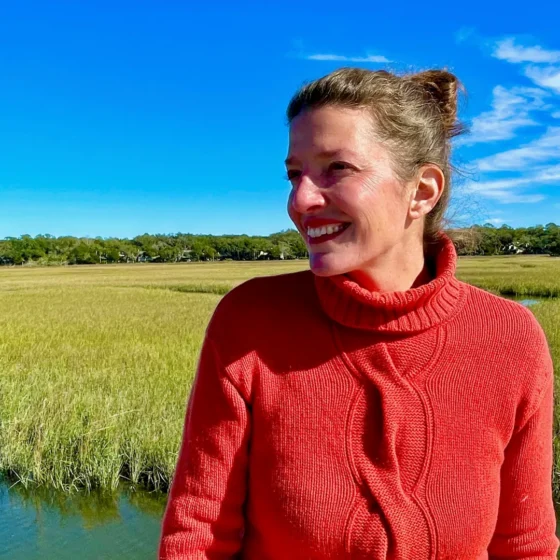 A woman with straight, brown hair in a bun, smiling and looking to the left. She stands in a watery field against a blue sky, and wears a red, turtleneck sweater.