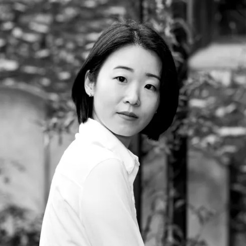 A grayscale photo of a woman with straight, black hair in a bob, and dark eyes. She sits outside, her torso faces the left while she looks forward, and she wears a white, collared shirt.