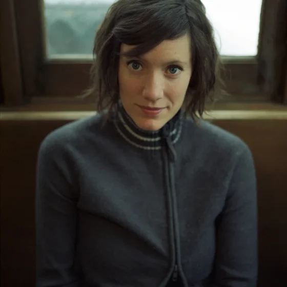 A woman with straight, brown hair and bangs to her chin, smiling. She sits in front of a window on a brown, wooden wall, and wears a gray sweater.