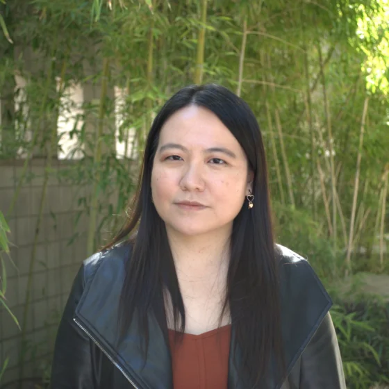A woman with long, straight, black hair that goes past her shoulders. She stands outside in front of a stone wall with bamboo growing in front of it. She wears a burnt-orange shirt underneath a black, leather jacket.