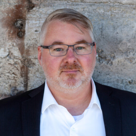 A man with gray hair and a beard standing in front of a stone background. He wears a white button-down with a black blazer over top.