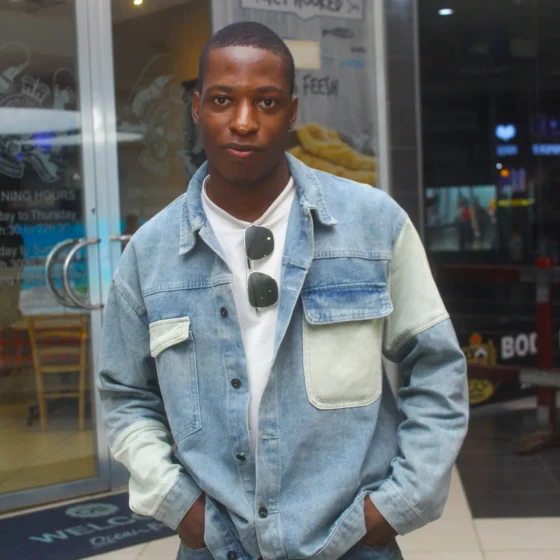 A Nigerian man with a buzz cut. He is standing in front of a store, putting his hands into the pockets of the white-and-blue, jean jacket he's wearing over top of a white shirt and blue jeans. A pair of square-shaped sunglasses hang from his collar.