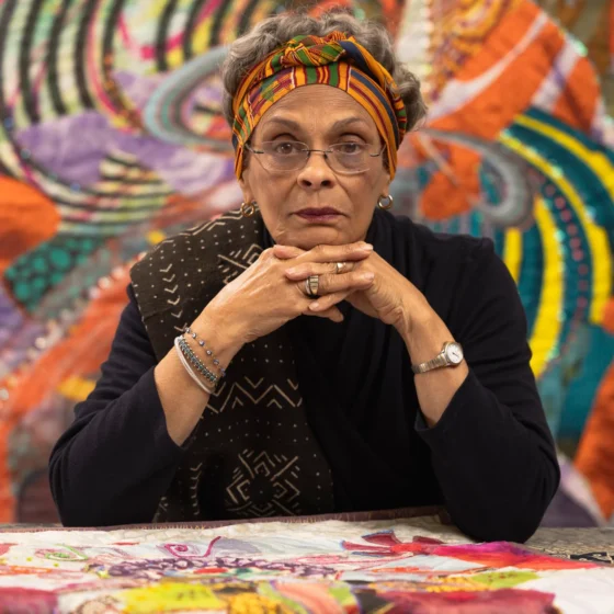 A woman with short, gray hair, resting her chin on clasped hands and looking forward. She sits before a table with a colorful tablecloth with intricate patterns, and in front of an equally colorful and patterned wall. She wears a colorful headband, rectangular glasses, and a black, long-sleeved blouse.