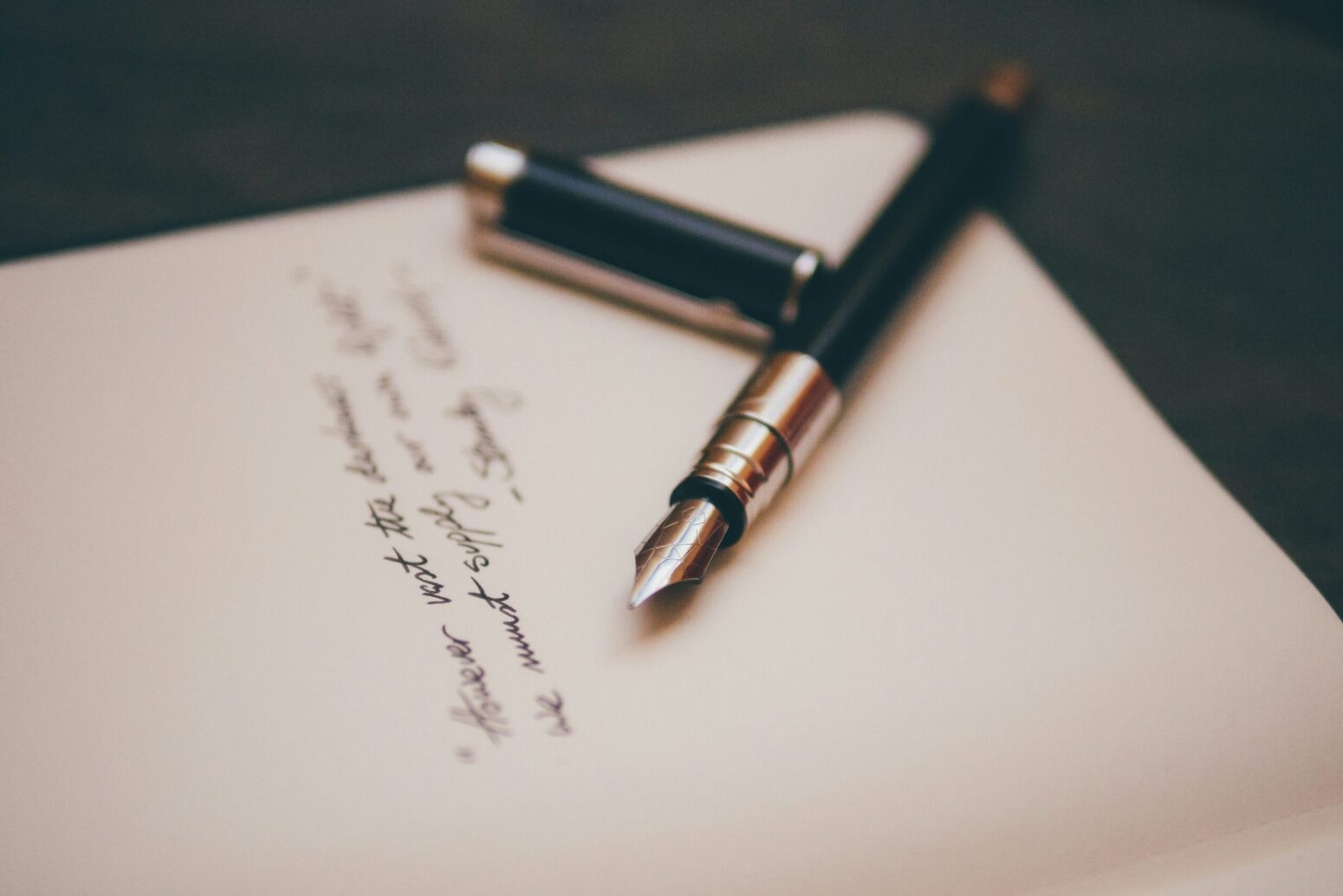 A paper sits on a desk with a black, calligraphy pen sitting, uncapped, on top of it. A a blurred sentence is written on the paper in cursive.