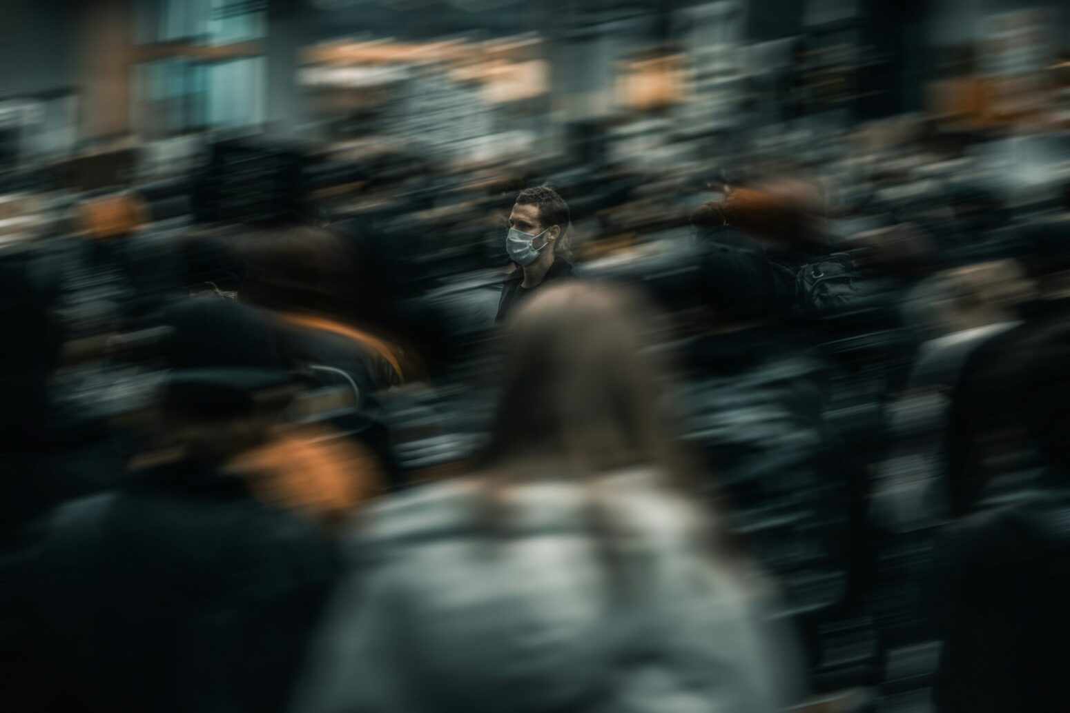 A man wearing a medical mask walking to the left in a crowd of people. All the people around him are blurred.
