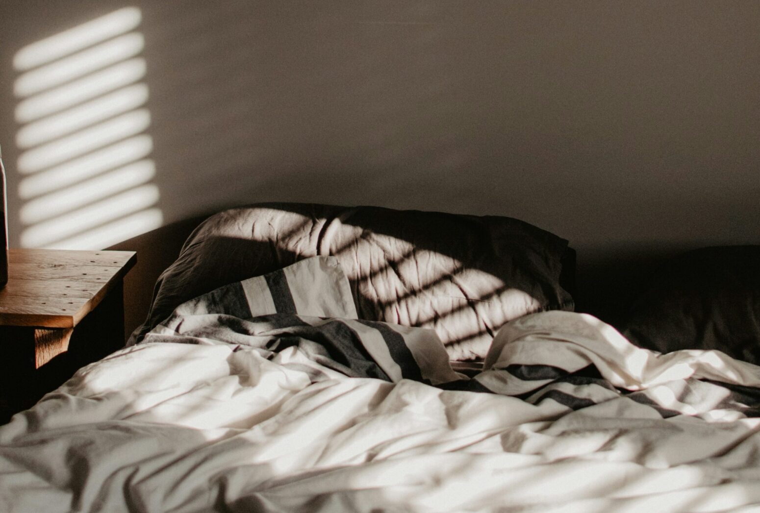 The sun shines through the blinds of a window onto a bed with white sheets, and gray pillows and blankets. The bed is up against a beige wall.