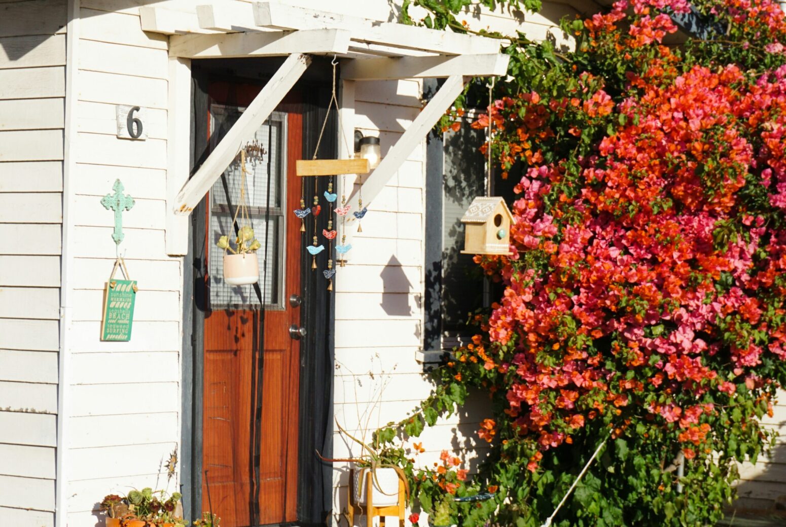 The front of a small, white house with a brown door and a large flower bush growing outside. The porch is small, with many potted plants sitting around it. A birdhouse, windchimes, and another plant are hanging from the small awning over top. The side wall of the house has a cross, and is labelled 6.