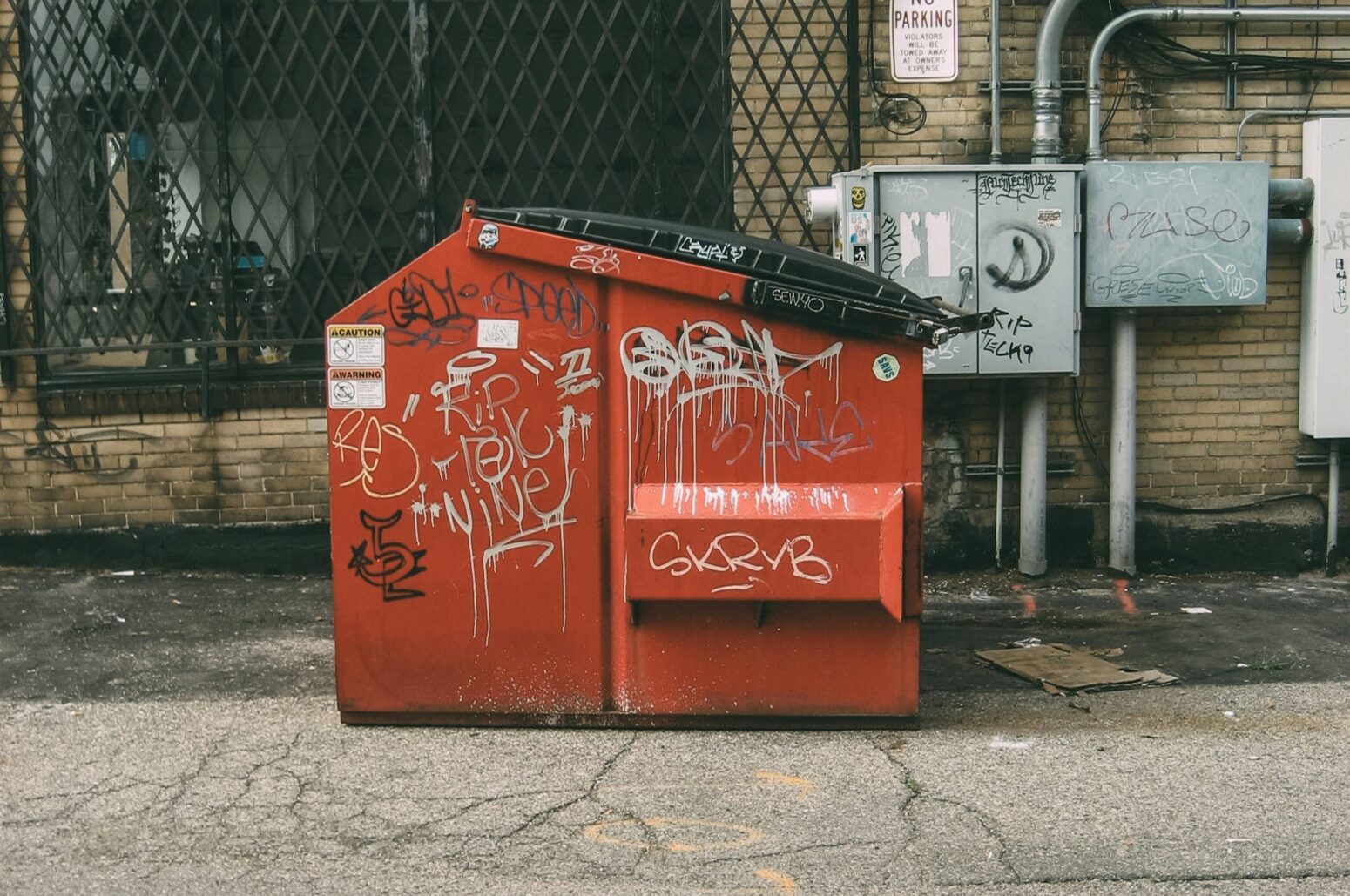 A large, orange dumpster with graffiti on the side. It sits on old asphalt in front of the electrical boxes of an old, brick building with a window covered by lattice, iron bars.
