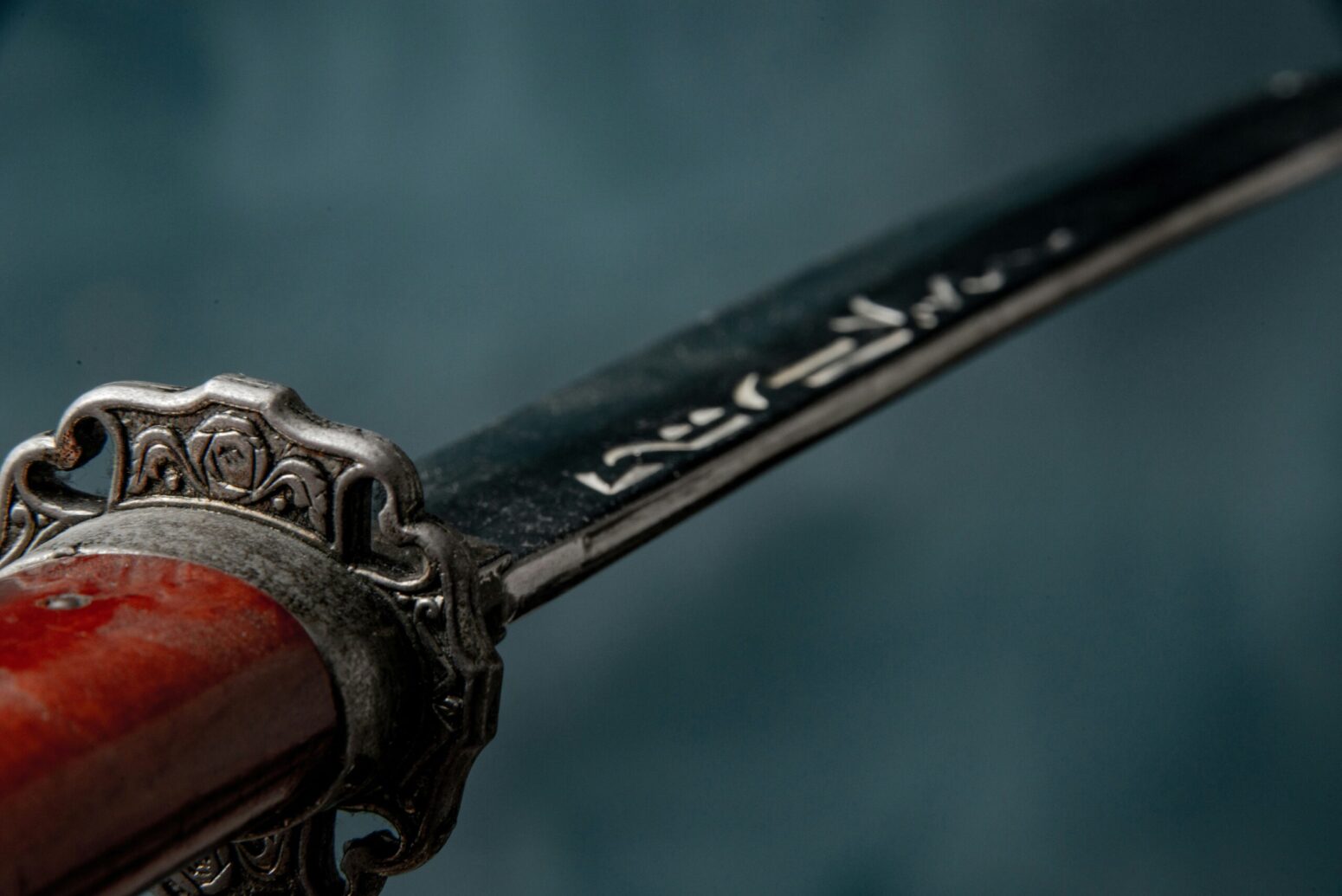 A close-up on the hilt of a sword, with a wooden handle and a intricate hand guard. The blade is pointing toward the top-right, and their are some markings near the base of it. The background is gray-blue.