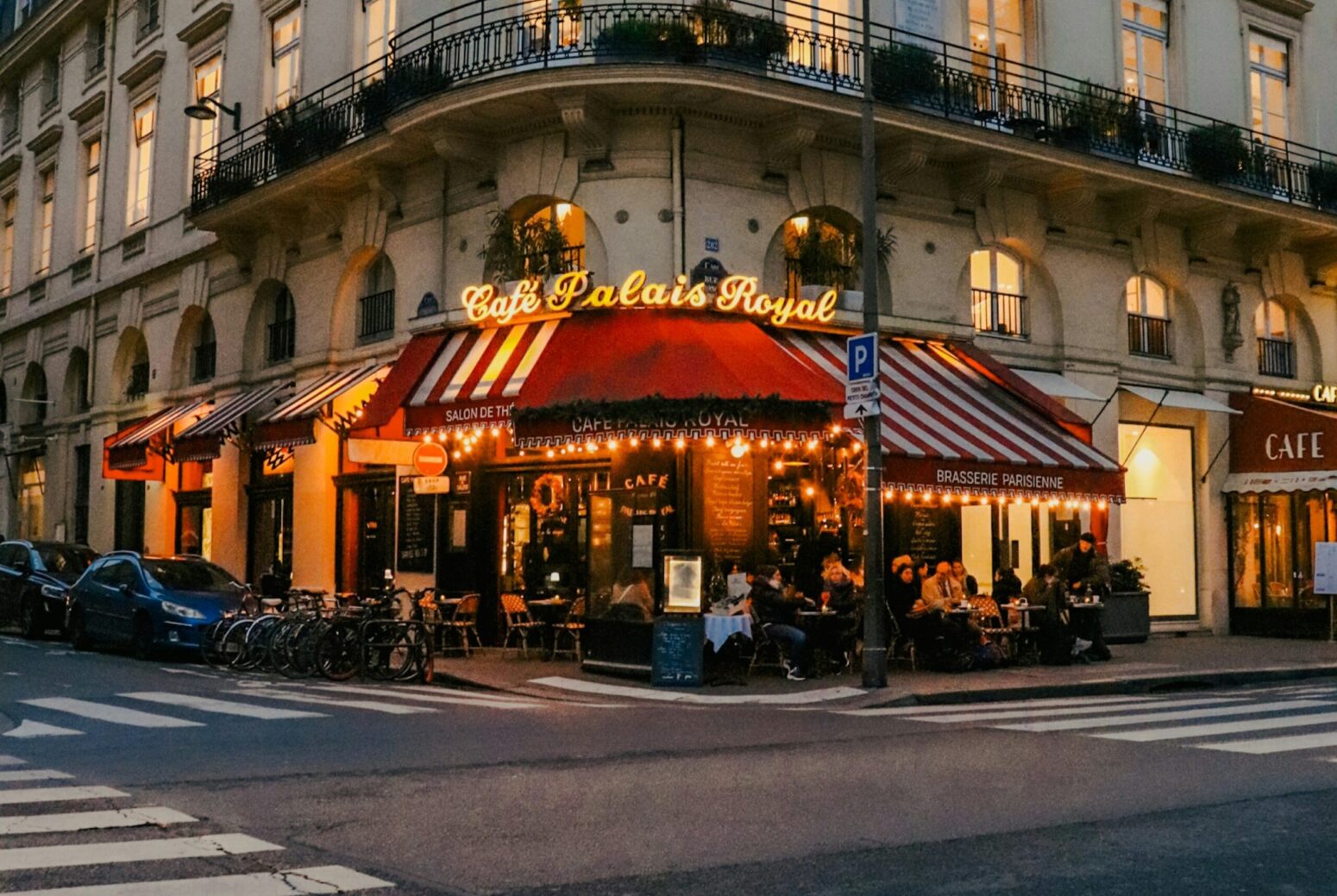The outside of Cafe Palais Royale in Paris, France. It sits on the corner of two streets, with outdoor seating covered by red canopies and illuminated with lights. The restaurant is the first floor of a larger, white building with lots of windows and a gray roof.