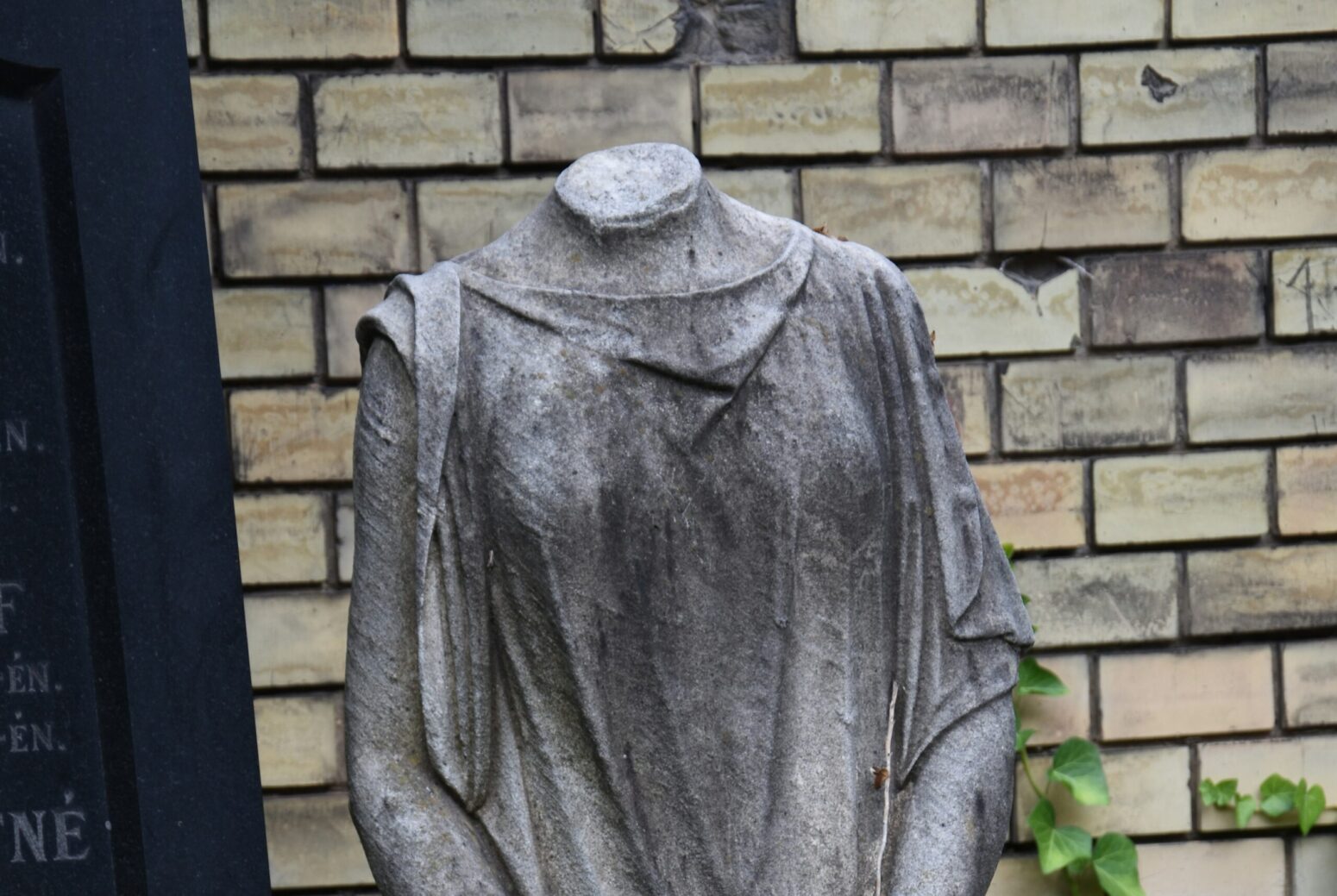 An old statue of a woman with its head missing. It stands in front of a wall of white bricks.