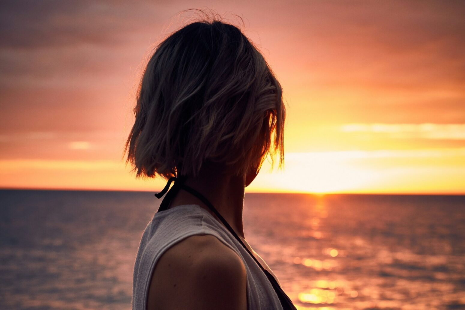 A woman with short, blonde hair, with her back facing the camera. She looks out at the horizon of the ocean during sunset. She wears a short-sleeved, white shirt.