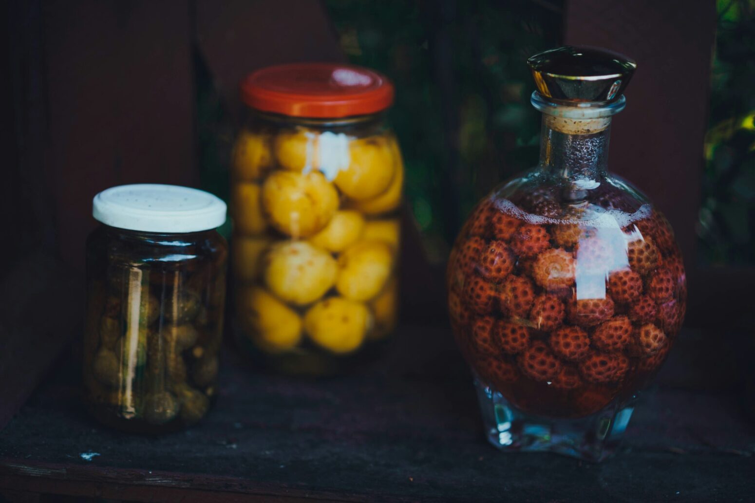 Three jars sit in front of a black background. On the right is a round jar full of liquid and Banksia nuts. In the middle is a jar with a red lid, filled with liquid and small potatoes. On the left is a small jar with a white lid, filled with pickles.