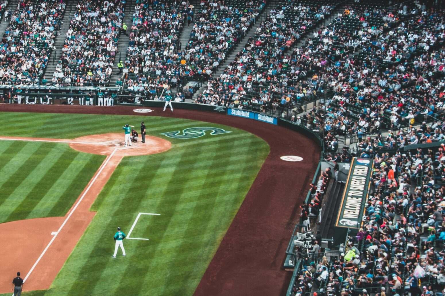 A view of a ballpark from a midsection on the side. The picture is of home plate. The stands are filled with people cheering for the Seattle Mariners.