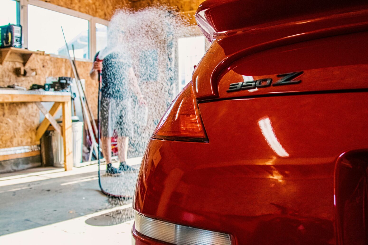 A man in a garage spraying down a red car with a hose. The walls are made of chipboard, with windows and a table with tools on it inside. The car's submodel reads "350 Z."