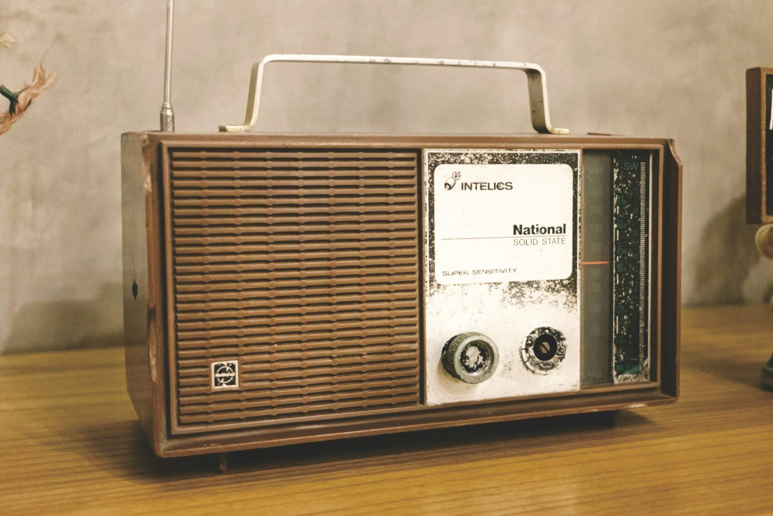 An old, electronic radio. It is brown with a metallic handle and a white interface. One of the two knobs is missing. It sits on a wooden desk in a room with white walls.