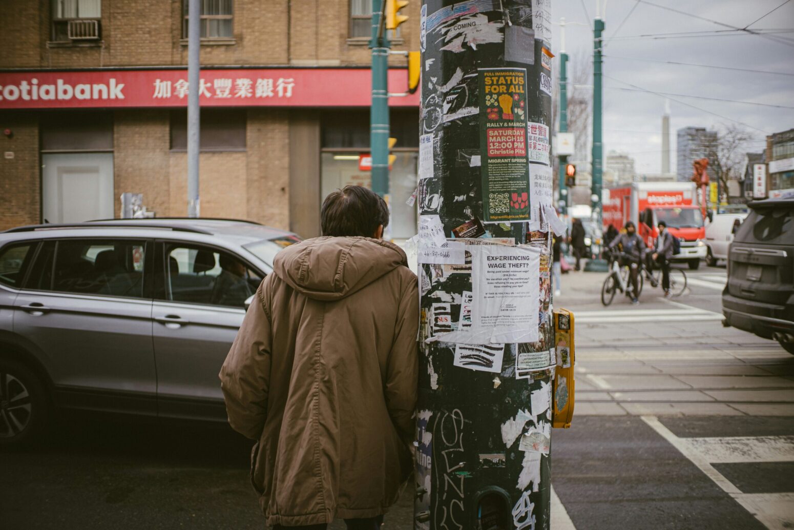 A man wearing a brown coat leans against a telephone pole in front of a crosswalk.. The pole is covered with old flyers. The background is of a city.