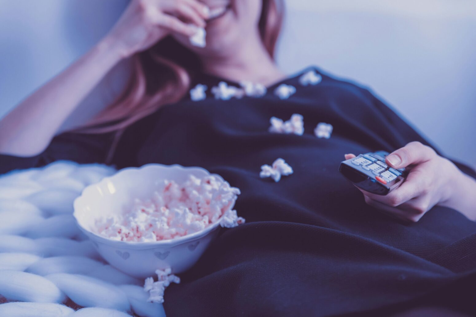 A woman with long, straight, blonde hair, laying back on a couch and smiling. She sits next to a bowl of popcorn that she is eating and that she has spilled all over her chest. She holds a TV remote in her left hand.