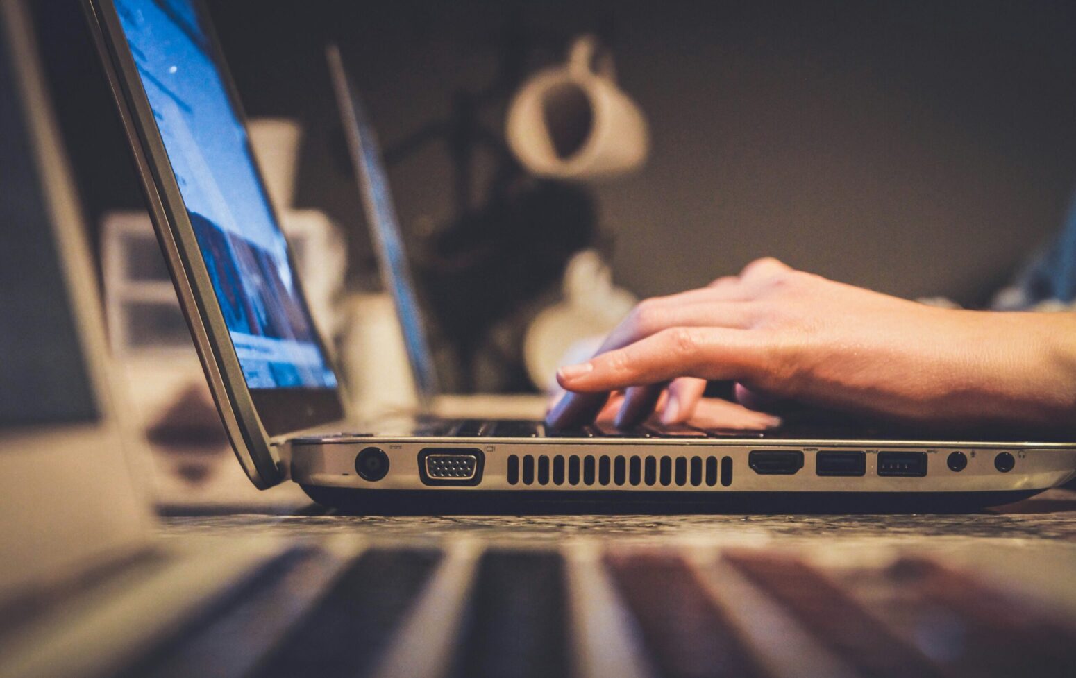 A side view of a person's hands, typing at a silver laptop on a white desk. In the background we can see a mug tree and a gray wall.