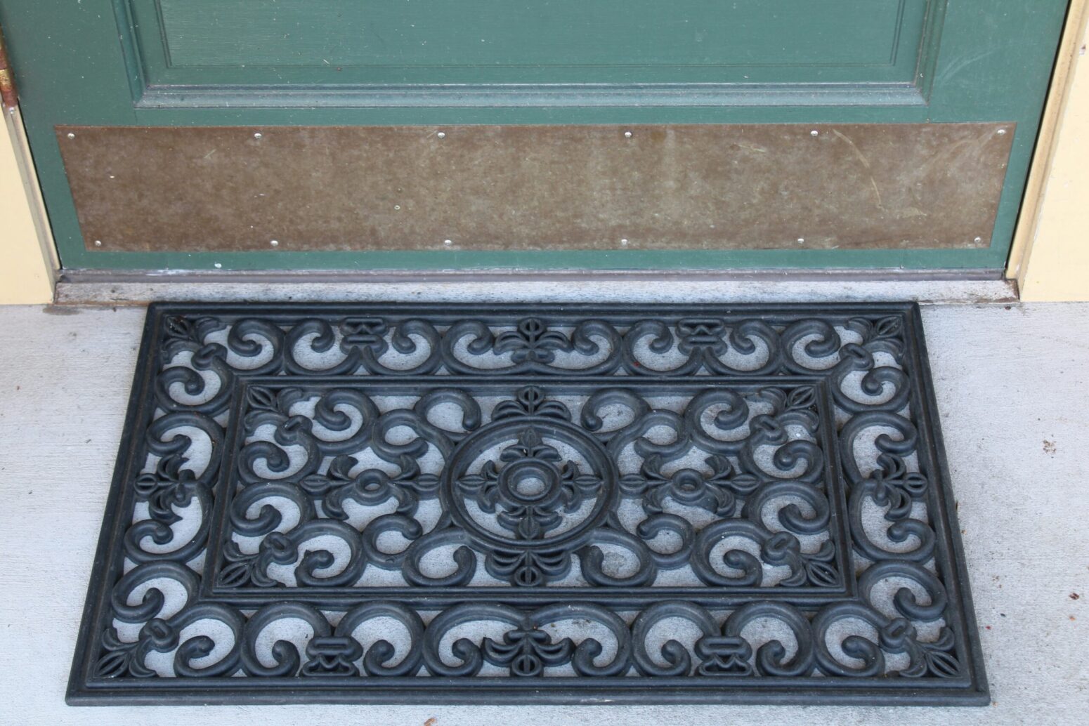 A black, rubber doormat on a gray, concrete porch. The porch is off of a light-yellow house with a green door.