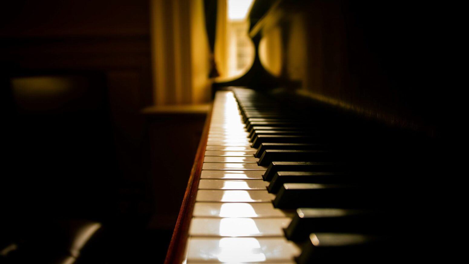 A side view a piano. It's keys are illuminated by the sun leaking through two yellow curtains that are mostly covering a window in the background.