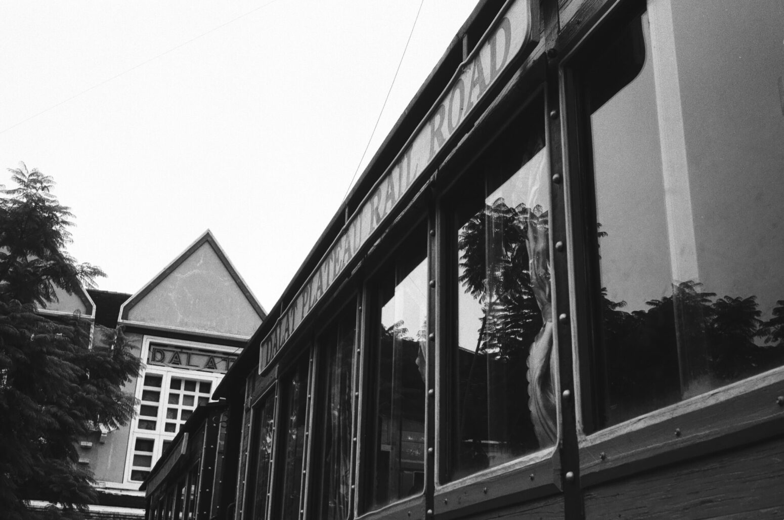 A grayscale photo of a train, stopped at a station. Above the windows reads "Dalat Plateau Rail Road." The station behind the train reads "Dalat."