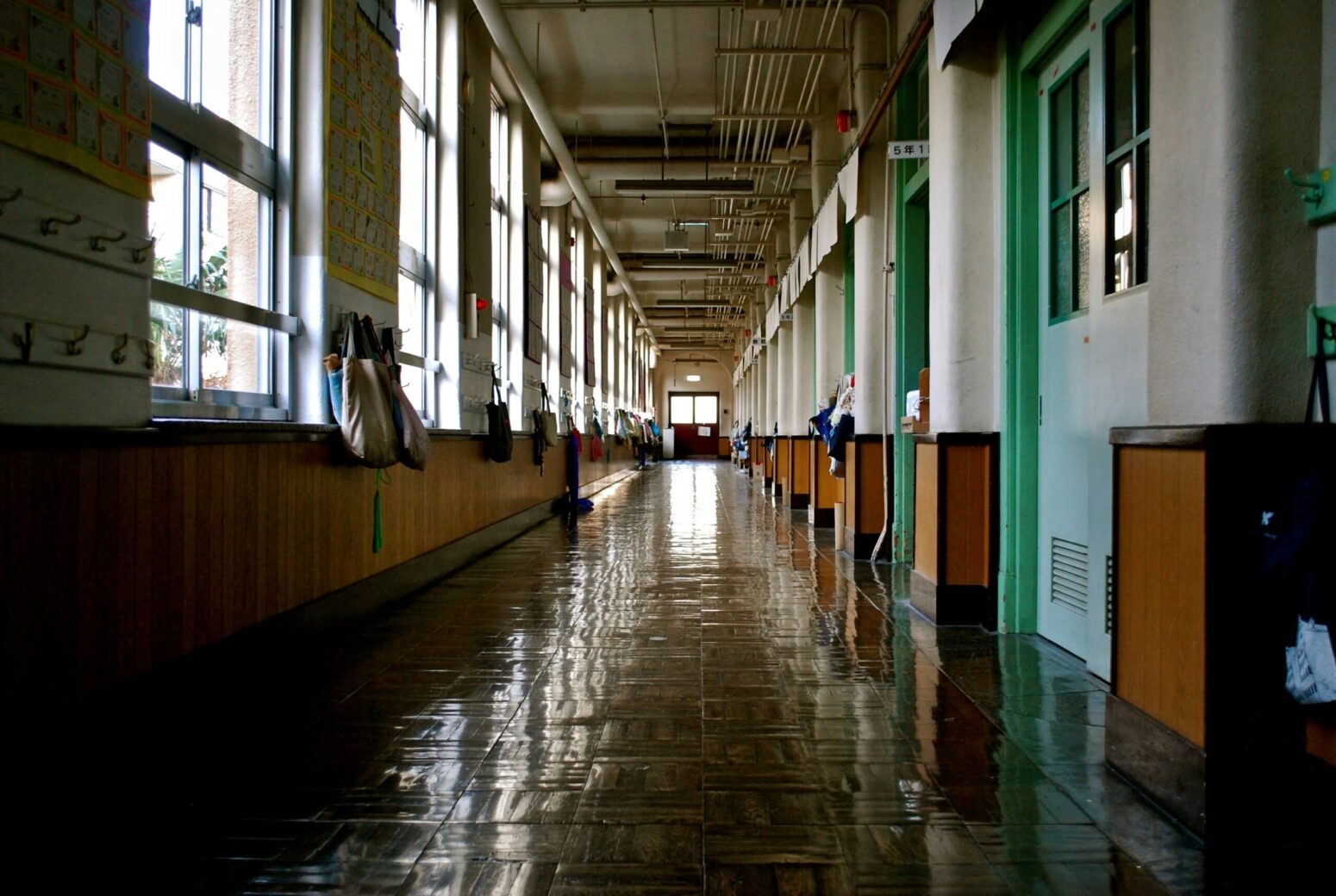A school hallway with brown, tile floors and half wooden, half white-painted walls. Windows line the left wall, doors to classrooms line the right. In between each window are hooks to hang tote bags onto. At the end of the hallway is a double-door.