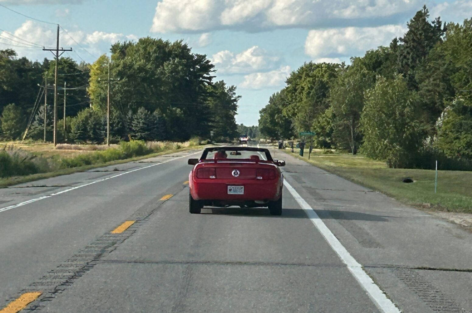 A red convertible, driving down a straight, country road. Powerlines and trees surround the road on either side.