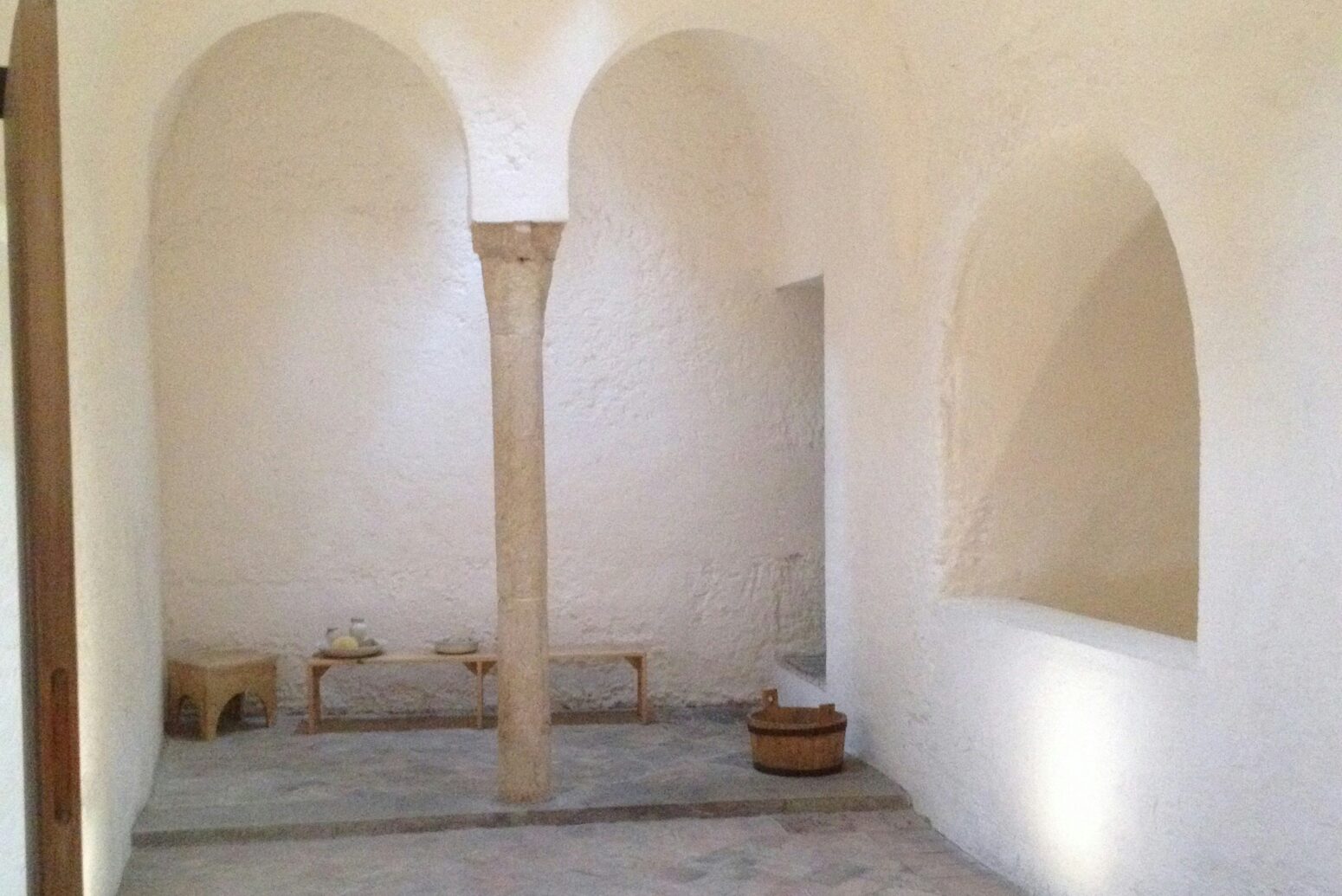 A room with white walls and stone floors. On the back wall is two archways, behind which sits a bench, a stool, and a bucket. On the right wall is a niche.