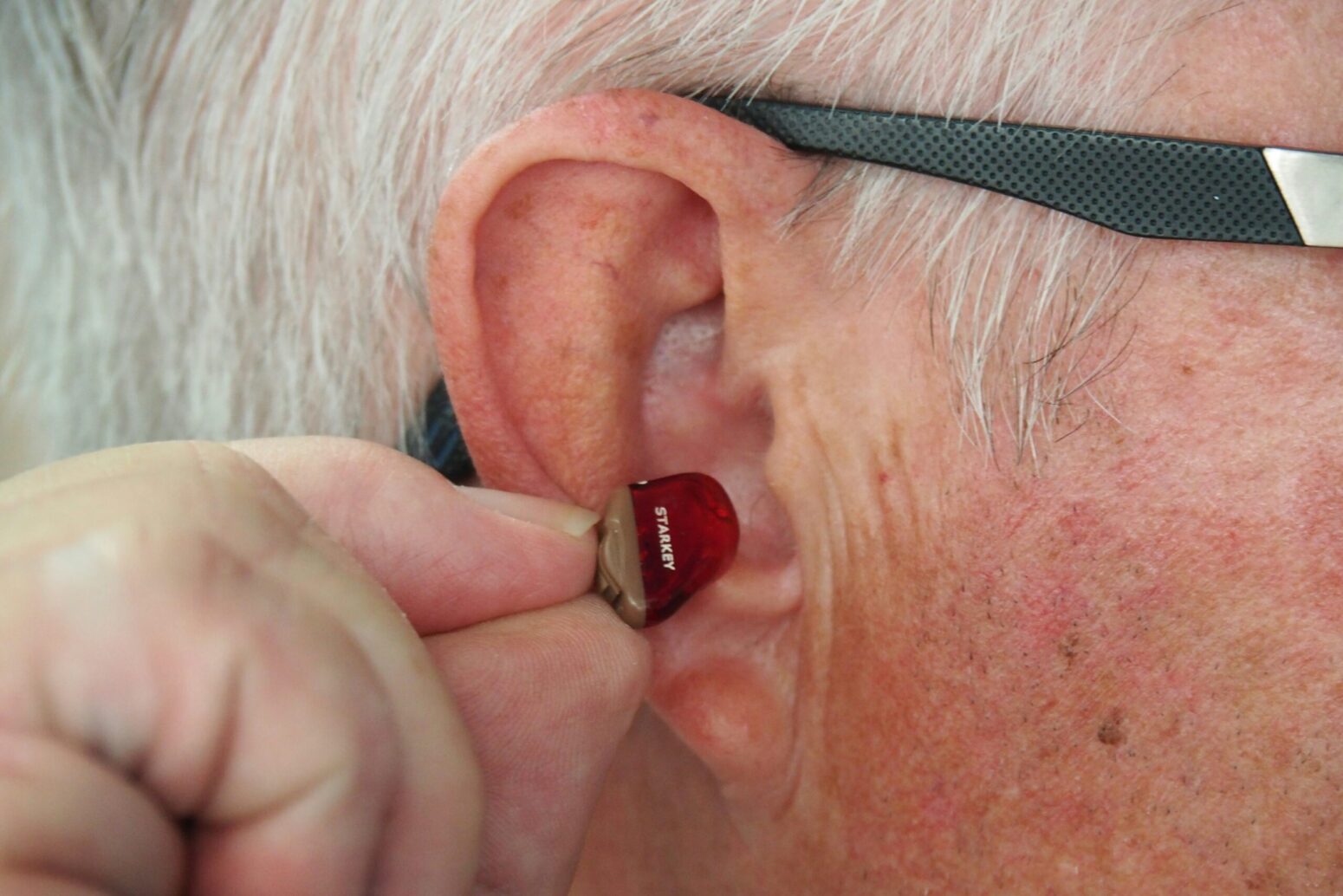 A close-up on an old man's right ear. He holds a small hearing aid up to it.