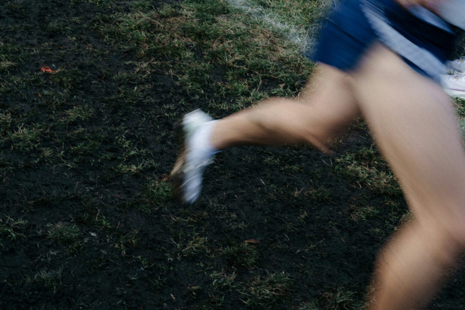 A blurry photo of someone's legs, running. They are running on grass and wearing navy shorts and white shoes.