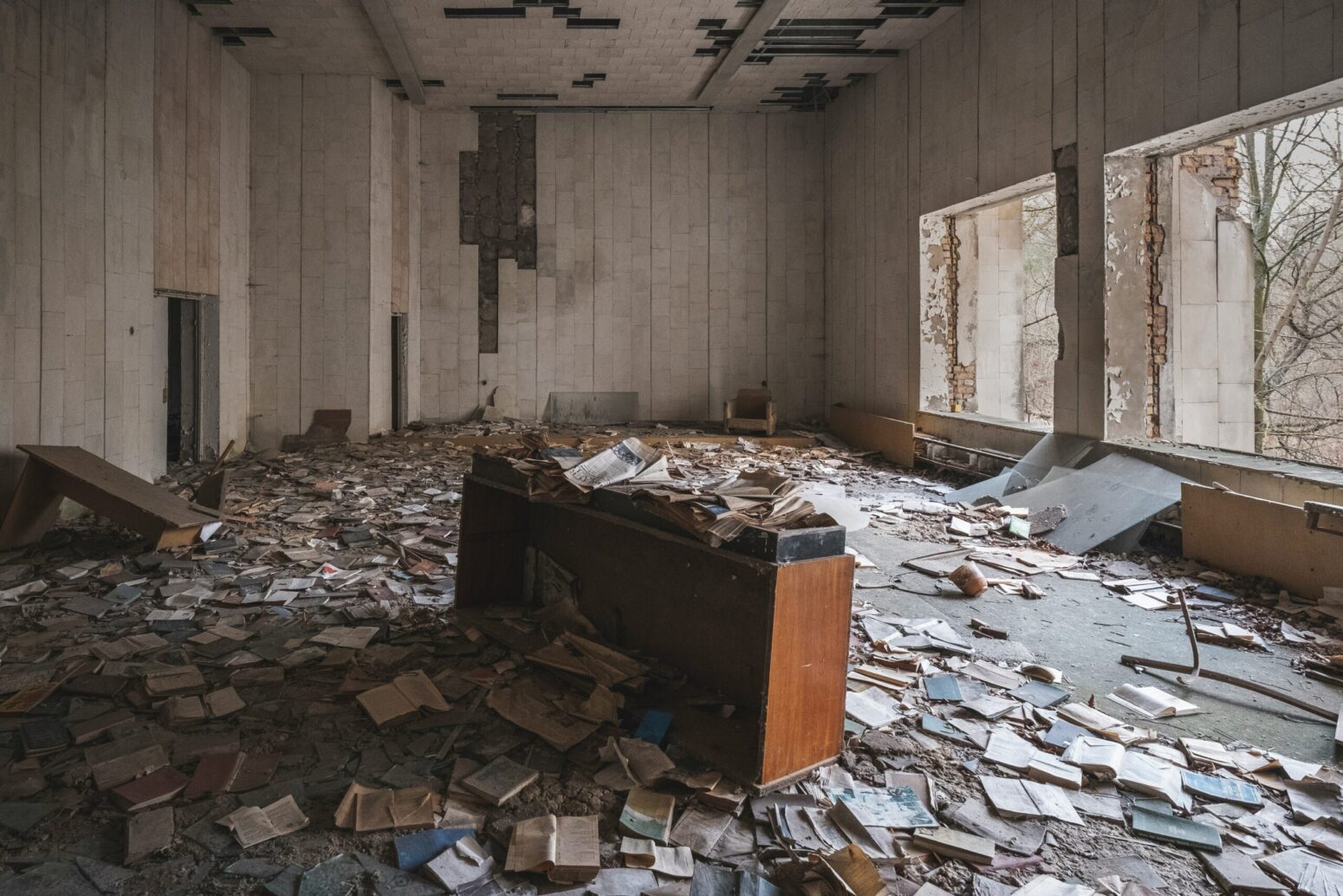 The inside of an abandoned building. The walls are white, and the panels are falling off. Where two windows once were on the right wall are now just two, square openings. Dirty, old books are littered all over the concrete floor, alongside a few broken, wooden desks.