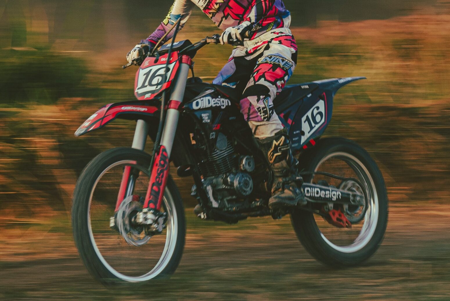 A person riding a dirt bike down a blurred, grassy hill. The dirt bike is red and white, and the person riding is wearing a red and white motocross gear to match.
