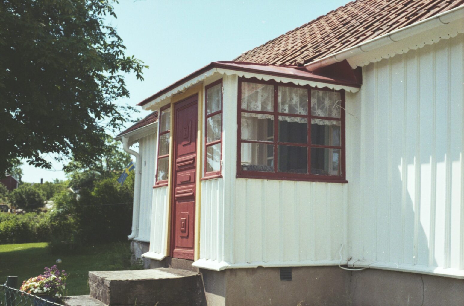 A small, white house with concrete foundation. The door, roof, and trimmings are painted red. In the background are some trees and a small flower bush.