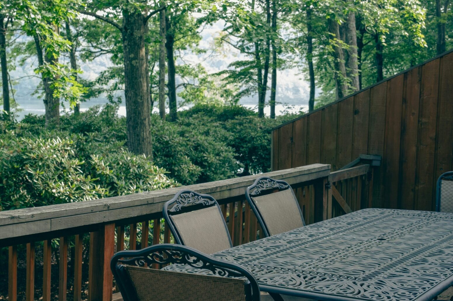 An ornate, metal table sits on a deck with four visible chairs surrounding it. The deck is surrounded by forest, with a lake in the background.