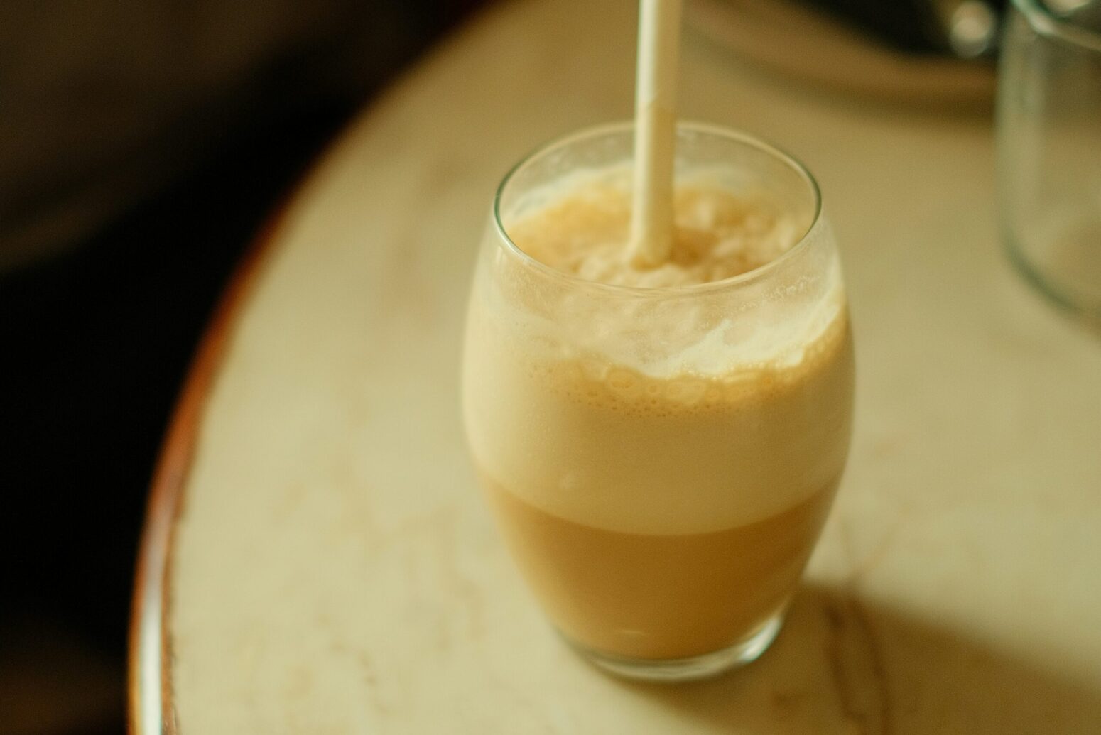 A cold, foamy coffee drink in a glass with a white straw on a white table.