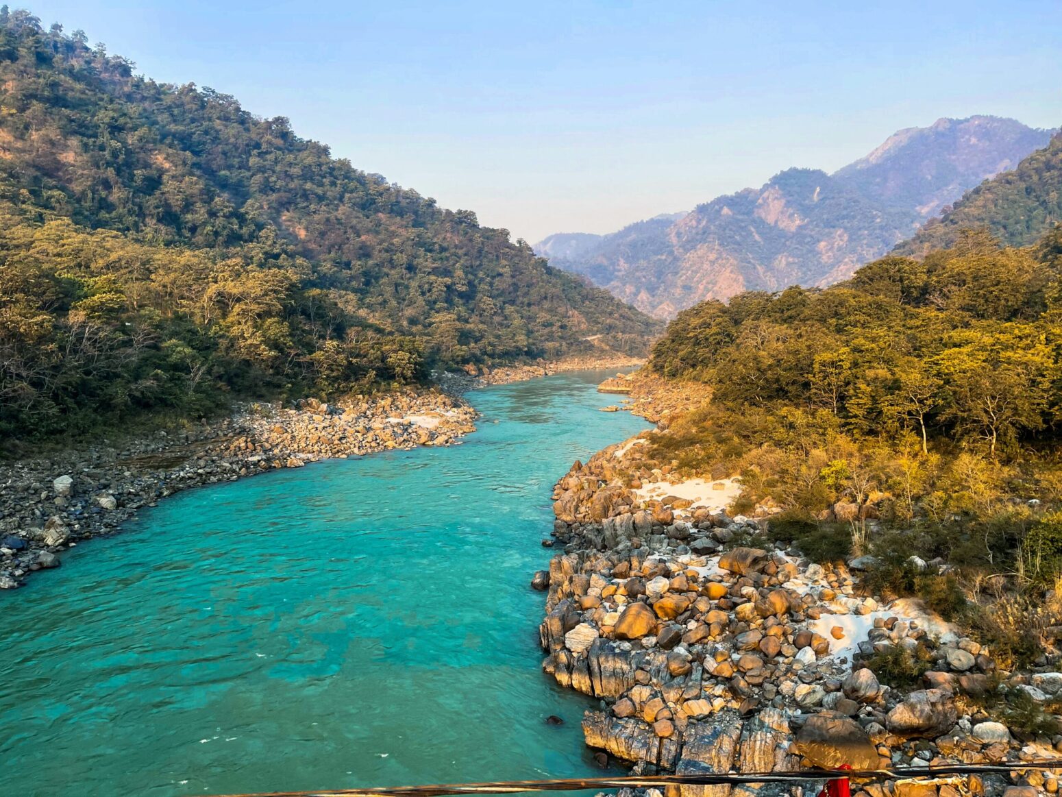 A picture of the Ganga river, a holy river that runs through India and Bangladesh. It flows upward, with rocks on either side and forest-covered mountains over the background.