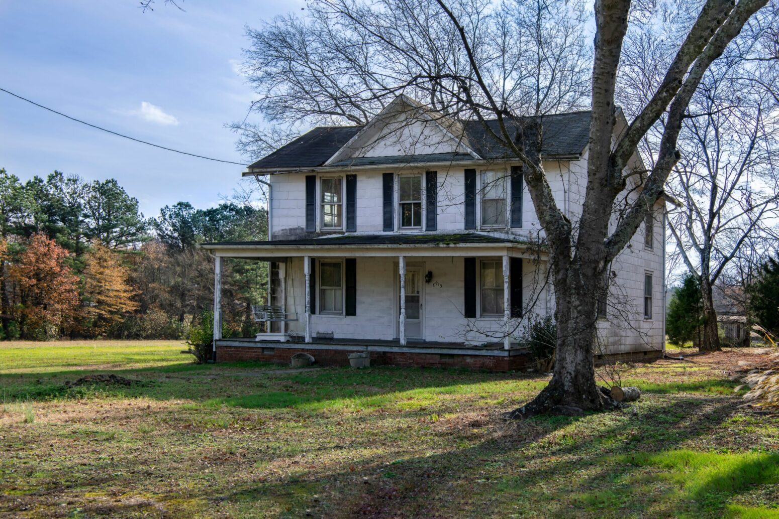An old, white, two-story house with a covered porch. The trim is black and the roof is gray. In the front yard there is a bare tree, and the rest of the house is surrounded by a clearing and woods further back.